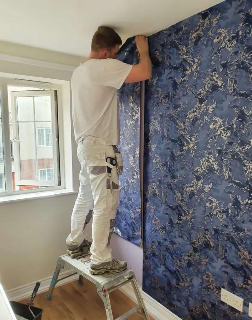 painters and decorators ruislip - Frazer stands on a ladder hanging blue and silver wallpaper