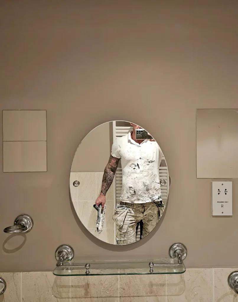 exterior house painting, kitchen cabinet painting, bathroom tile replacement - image shows beige bathroom wall with glass shelf under oval mirror - decorating contractor Frazer can be seen in the mirror wearing paint covered workwear